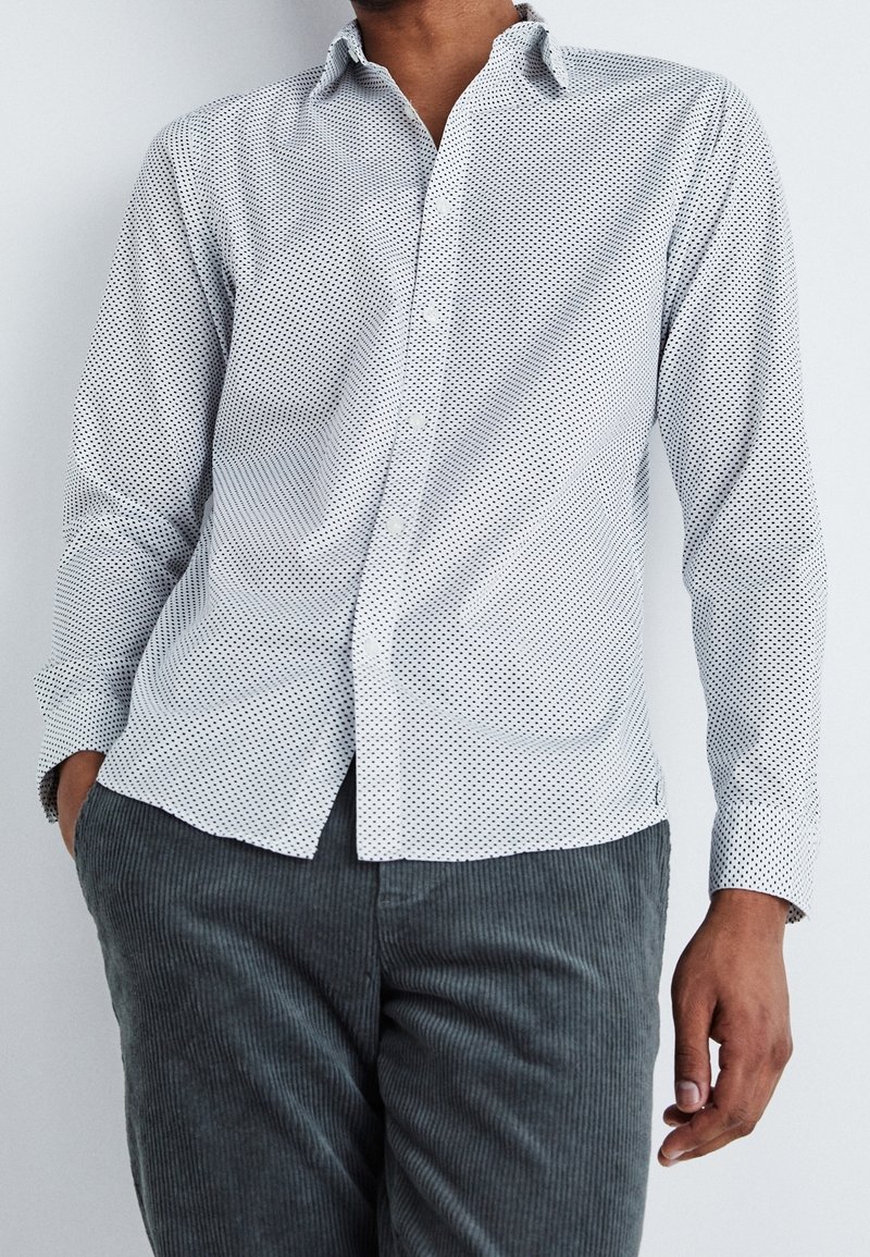 Camicia bianca con motivo a piccoli pois neri, maniche lunghe e colletto appuntito; abbinata a pantaloni in velluto a coste grigi testurizzati.