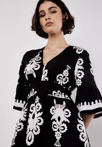 Une femme aux cheveux courts pose avec confiance, vêtue d'une robe noire ornée de motifs floraux blancs audacieux et de larges manches.