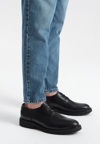 Scarpe in pelle nera con una finitura liscia, punta rotonda e design con lacci, abbinate a jeans di denim azzurro chiaro, leggermente svasati alle caviglie.
