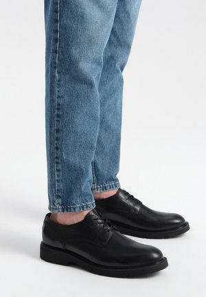 Zwarte leren schoenen met een gladde afwerking, ronde neus en vetersluiting, gecombineerd met lichtblauwe denim jeans, die iets taps toelopen bij de enkels.