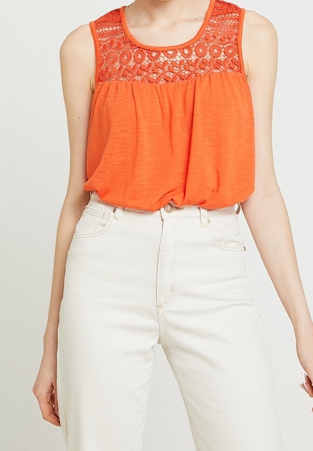 Pomarańczowy bezrękawnik z koronkowymi detalami przy dekolcie, zwiewna tkanina i teksturowany gorset, zestawiony z białymi spodniami z wysokim stanem.