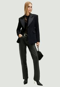 Zwarte blazer met satijnen kleppen, doorschijnend zwart topje, zwarte leren broek, puntige hakken en een gestructureerde zwarte handtas.