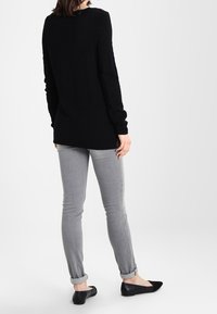 Woman standing, wearing a black long-sleeve sweater, light gray rolled-up skinny jeans, and black flat shoes on a white background.