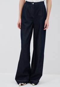 Tummansiniset korkeavyötäröiset väljät farkut, joissa on etupillit, nappikiinnitys ja erottuva ompelu, esitelty vaalealla taustalla.