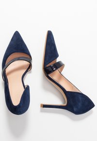 Coppia di scarpe blu navy in suede con tacco alto e punte appuntite, con cinturini in pelle verniciata nera su sfondo bianco.