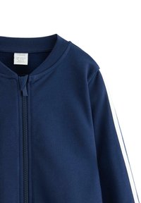 Navy blauwe kinderjas met ritssluiting aan de voorkant en een witte streep langs de mouw, geribbelde kraag, maatlabel zichtbaar aan de binnenkant.