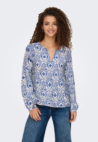 Blå och vit mönstrad blus med långa ärmar, design med V-ringning och en strukturerad tyg. Parat med blå denimjeans.