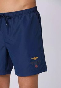 Pantaloni da bagno blu navy con cintura elastica e coulisse, caratterizzati da un logo ricamato e una patch con dettagli dorati.