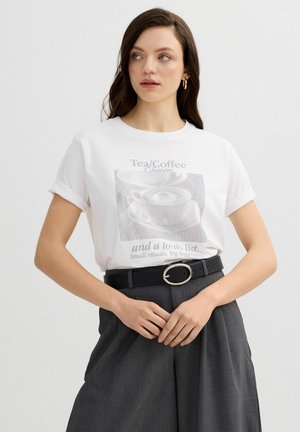 Mujer joven con cabello oscuro que lleva una camisa blanca de manga corta con estampado y pantalones grises de talle alto, manos cruzadas en la cintura, mirando de lado.