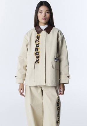 Młoda kobieta ubrana w beżową kurtkę i spodnie z ciemnobrązowymi kwiatowymi wzorami oraz brązowym kołnierzem, stojąca na białym tle.