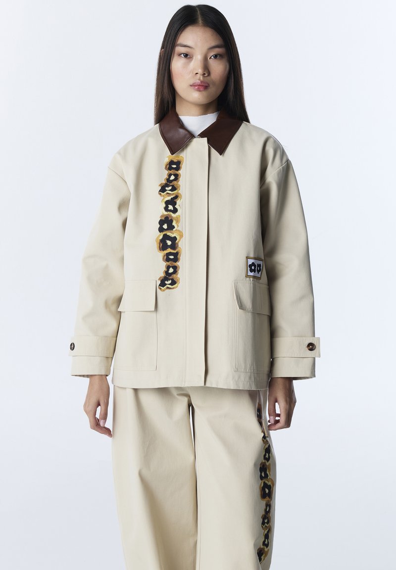 Jonge vrouw die een beige jas en broek draagt met donkerbruine bloemmotieven en een bruine kraag, staand tegen een witte achtergrond.