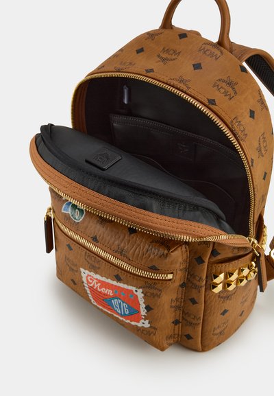 Sac à dos en cuir synthétique marron avec intérieur noir, fermeture à glissière et logo MCM. Il comprend des clous, un patch et plusieurs compartiments pour une meilleure organisation.
