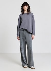 Donna con capelli ricci che indossa una felpa grigia larga, pantaloni grigi a gamba larga con cordoncino e sneaker bianche, in piedi su uno sfondo neutro.