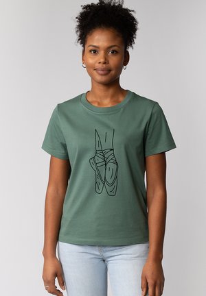 Femme portant un t-shirt vert avec un dessin au trait noir de chaussures de danse classique et un jean bleu clair, debout devant un fond gris uni.