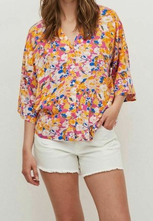Kvinde iført en løs, farverig blomstret bluse med pink, orange, blå og hvide accenter kombineret med hvide shorts med frynset kant.