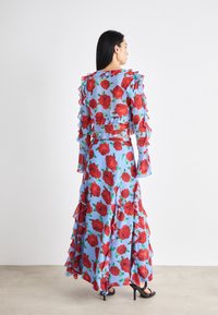 Maxi dress in light blue fabric with red rose print, featuring ruffled sleeves and hem. The design has a fitted bodice with a flowing skirt.