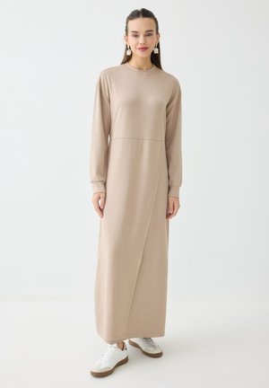 Kvinde iført lang beige kjole med lange ærmer og hvide sneakers står mod en hvid baggrund.