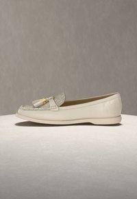 Μπεζ δερμάτινο μοκασίνι με ανάγλυφο ύφασμα στην επάνω πλευρά, χρυσούς μεταλλικούς λεπτομέρειες και διπλές φούντες, τοποθετημένο πάνω σε ουδέτερη επιφάνεια.