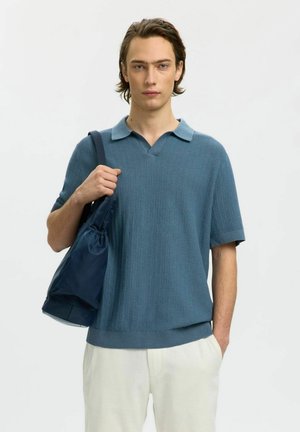 Jonge man die een gebreid blauw poloshirt en een witte broek draagt, met een donkerblauwe schoudertas over zijn linkerschouder.