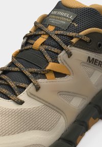 Ποδηλατικό παπούτσι Merrell σε μπεζ και μαύρο με κίτρινες λεπτομέρειες, λεπτομερές επάνω μέρος από δίχτυ, κορδόνια με σχέδιο και ανθεκτική σόλα σχεδιασμένη για υπαίθρια χρήση.