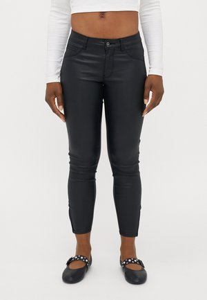 Kobieta w czarnych, wysokich, dopasowanych spodniach z sztucznej skóry, białym, długim crop topie i czarnych balerinkach Mary Jane z ćwiekami.