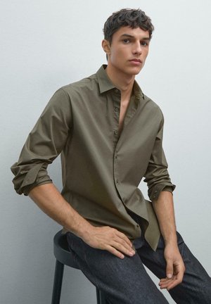 Olive green button-up shirt with a classic collar, rolled sleeves, and a smooth texture, paired with dark jeans, seated on a black stool.