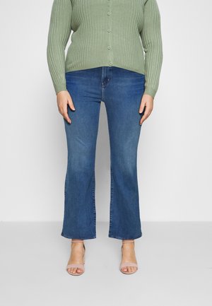 Persoon die blauwe jeans met rechte pijpen draagt, een lichtgroene geribde gebreide trui met knopen en beige open teen hakken, staand tegen een effen achtergrond.