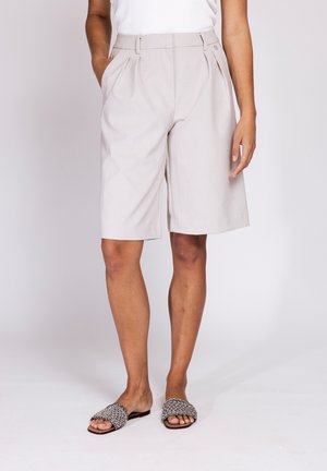 Person wearing light beige knee-length tailored shorts, white top, and patterned open-toe slip-on sandals, standing against white background.