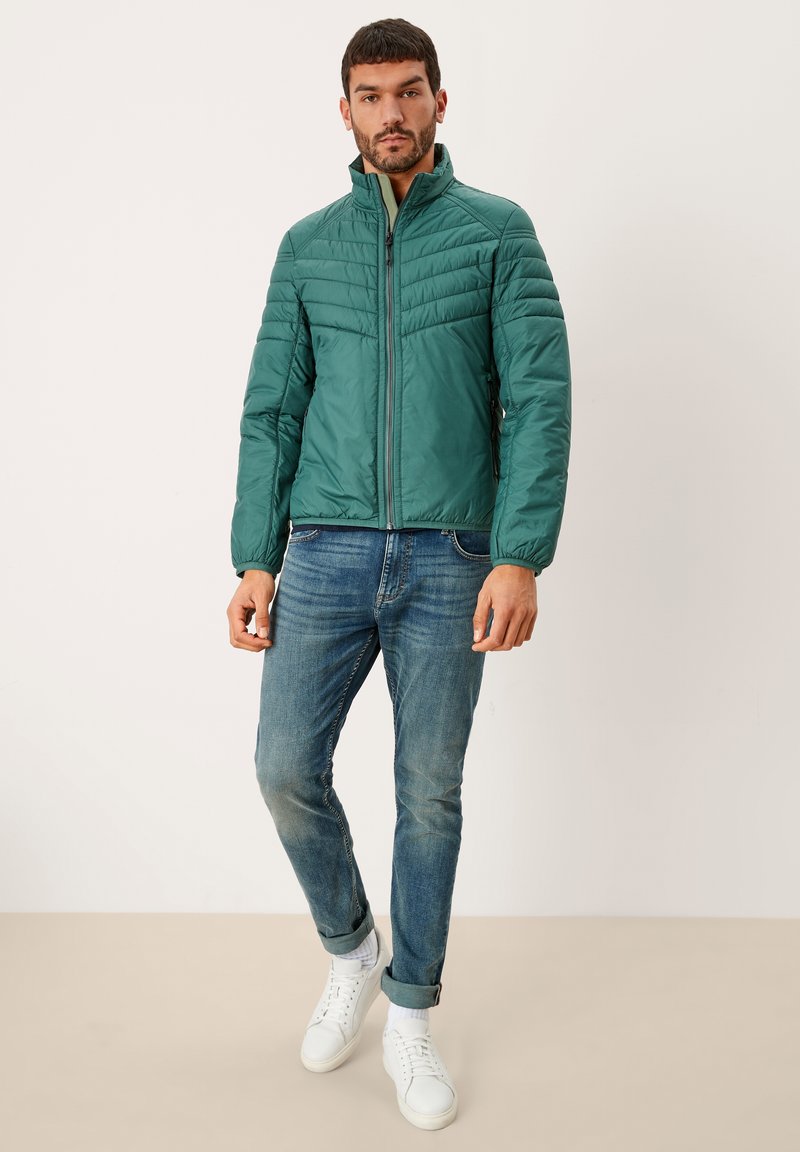 Giacca imbottita verde con colletto alto, dotata di chiusura a zip e polsini a costine. Abbinata a jeans blu e sneakers bianche.