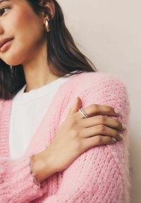 Vaaleanpunainen neulecardigan, jossa on pörröinen pinta, on puettu valkoisen paidan päälle. Käsi, jossa on monivärinen rengas, lepäilee hihalla.