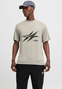 Lys beige kortærmet t-shirt med et stort sort grafisk logo på forsiden, parret med sorte træningsbukser. Glat tekstur.