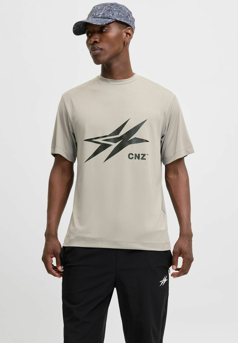 Lys beige kortærmet t-shirt med et stort sort grafisk logo på forsiden, parret med sorte træningsbukser. Glat tekstur.
