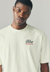 Tricou din bumbac verde deschis, cu un logo negru "Ford" și text roșu "BRONCO" pe partea stângă a pieptului. Mâneci scurte, guler rotund.