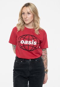 Red cotton t-shirt featuring a black globe design with the word "oasis" in bold white letters. Paired with black high-waisted jeans.