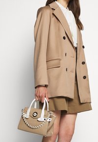 Beige tuplakietė švarkas su juodais mygtukais, derinamas su rudos spalvos šortais. Modelis laiko tekstūruotą beige rankinę su baltais rankenėlėmis ir grandine.