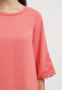 Korálový top s kulatým výstřihem a rozšířenými rukávy, zdobený jemnou krajkovou aplikací na koncích rukávů a hladkou texturou látky.