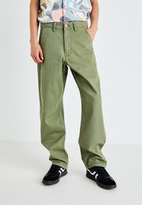 Grüne, locker sitzende Hose aus Baumwolle. Verfügt über vordere Taschen, einen standardmäßigen Reißverschluss und eine leichte Waschung. Trägt schwarze Sneakers.