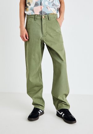 Grüne, locker sitzende Hose aus Baumwolle. Verfügt über vordere Taschen, einen standardmäßigen Reißverschluss und eine leichte Waschung. Trägt schwarze Sneakers.