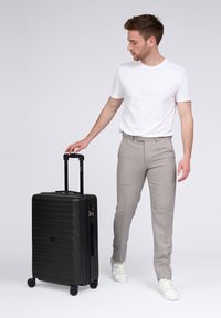 Junger Mann in weißem T-Shirt und grauen Hosen, der den Griff eines schwarzen rollenden Koffers auf weißem Hintergrund hält.