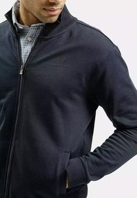 Ciemnoniebieska kurtka zapinana na zamek z miękkiego materiału, z wysokim kołnierzem, bocznymi kieszeniami i subtelnym logo na klatce piersiowej.