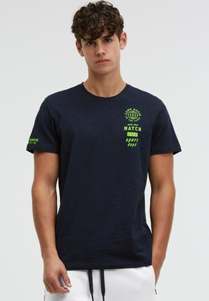 Junger Mann mit lockigem Haar, der ein dunkelblaues T-Shirt mit neongrünem Sporttext und weißen Shorts vor einem schlichten Hintergrund trägt.