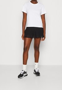 Žena oblečená v bílém tričku s krátkým rukávem, černé plisované mini sukni, bílých ponožkách a černobílých teniskách Nike stojí na jednobarevném pozadí.