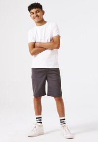 Camiseta de algodón blanca, pantalones cortos grises, zapatillas blancas, calcetines con patrón en blanco y negro. El modelo está de pie con los brazos cruzados, sonriendo.