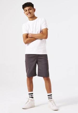 T-shirt en coton blanc, shorts gris, baskets blanches, chaussettes à motifs noir et blanc. Le modèle se tient les bras croisés, souriant.