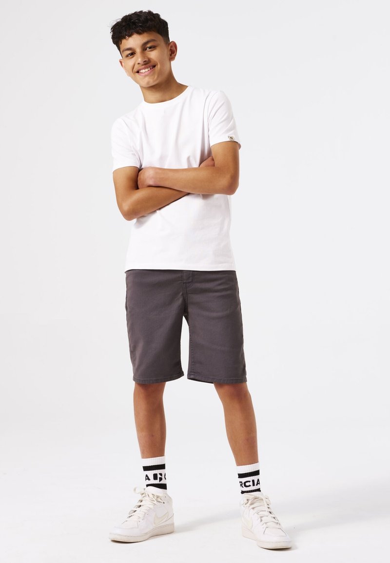 Camiseta de algodón blanca, pantalones cortos grises, zapatillas blancas, calcetines con patrón en blanco y negro. El modelo está de pie con los brazos cruzados, sonriendo.