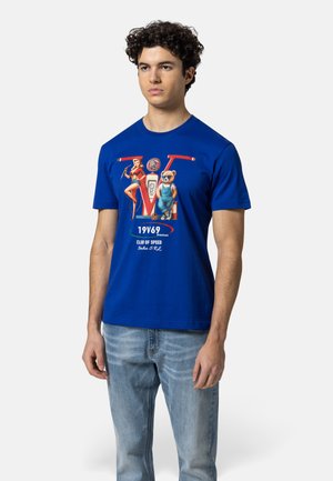 Junger Mann mit lockigem Haar, der ein blaues T-Shirt mit Aufdruck trägt, das eine Pin-up-Frau, einen Bären in Latzhose und den Schriftzug "19V69 Club of Speed" zeigt.