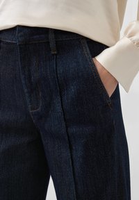 Ciemnoniebieskie dżinsy z delikatnym połyskiem, o prostym kroju nogawek, bocznych kieszeniach i kontrastowym pomarańczowym przeszyciu. Gładka tekstura.