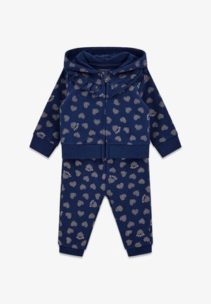 Tumesinine spordikomplekt, kus on lukuga kapuutsiga jakk ja püksid. Omab üle kogu pinna südame mustrit ja hõbedast teksti "GUESS". Pehme puuvillane materjal.