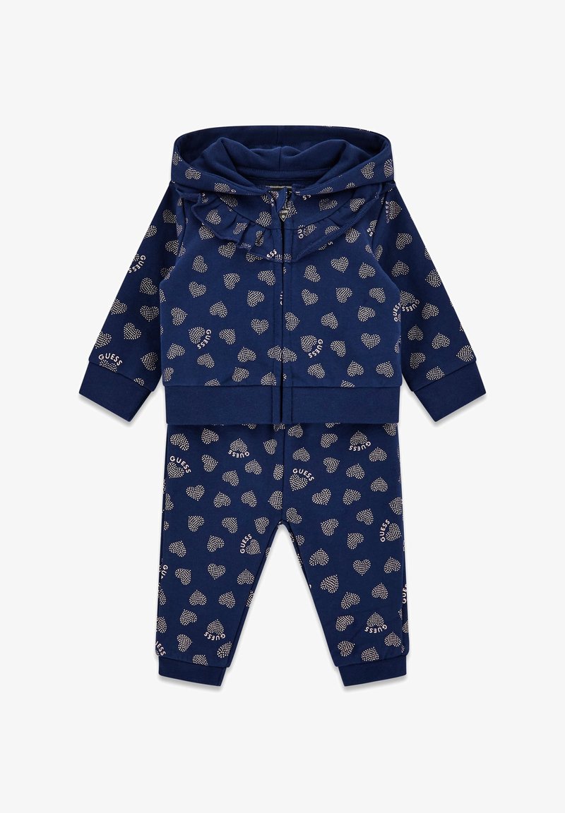 Navy tracksuit set with zip-up hooded jacket and pants. Features all-over heart pattern and "GUESS" text in silver. Soft cotton material.