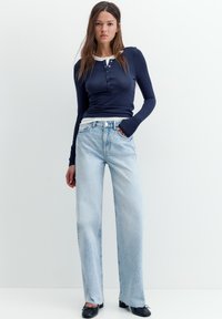 Femeie în picioare pe fundal alb, purtând un pulover bleumarin cu mânecă lungă, blugi cu talie înaltă de culoare albastru deschis și pantofi negri, cu o mână în buzunar.